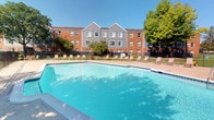 This outdoor amenity area centers around a large, rectangular swimming pool with clear, blue water and a classic geometric design featuring angled corners. The pool is equipped with multiple stainless steel entry rails and is surrounded by a spacious concrete deck that provides ample room for relaxation and movement. A row of lounge chairs lines one side of the pool deck, offering dedicated sunbathing space under open skies. The area is enclosed by a black metal fence, ensuring privacy and security, while mature deciduous trees frame the perimeter, providing natural shade and seasonal color.

An adjacent single-story brick building with a pitched roof serves as a poolside amenity structure. Inside, a shared laundry facility is equipped with commercial-grade top-loading washers and dryers, a folding table, and bright overhead lighting. A connecting hallway leads to individual storage units, each numbered and accessed via dedicated doors, providing organized and secure personal storage options. The hallway is well-lit with recessed lighting and leads to an exterior door with a windowed panel for added natural light. Together, these amenities complement the outdoor pool experience, offering practical conveniences within the same facility.