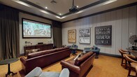 This purpose-built home theater room is thoughtfully designed to deliver a premier cinematic experience. A large wall-mounted projection screen anchors the front of the room, paired with a ceiling-mounted projector and a substantial dark wood media console below that houses audio-visual equipment. The tray ceiling adds architectural depth, framed by dark trim that complements the room's sophisticated palette, while recessed lighting provides controlled ambient illumination.

The seating arrangement is generously appointed with multiple full-size rich brown leather sofas and plush blue armchairs, all accented with faux fur throw pillows for added comfort. Warm golden-toned carpet covers the floor throughout, contributing to the room's inviting atmosphere. Floor-to-ceiling vertical board paneling in a neutral gray-beige tone lines the walls, lending both acoustic character and a refined aesthetic.

Adding a charming, vintage flair, framed cinema-style "General Admission" and "Admit One" posters decorate the side walls, evoking the nostalgia of classic movie houses. Decorative wall art, including circular metalwork pieces, further enriches the space. A dining or game table with chairs is tucked along one side wall, offering additional utility. Heavy drape curtains frame the screen wall, completing the theatrical ambiance of this well-appointed entertainment space.