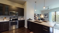 This single-floor residence showcases a thoughtfully designed open-concept layout that seamlessly connects the kitchen, dining, and living areas. The kitchen is a standout feature, outfitted with dark espresso cabinetry, a tile backsplash, and a full suite of stainless steel appliances including a French door refrigerator, electric range, built-in microwave, and dishwasher. A peninsula with a white quartz countertop and undermount sink doubles as a breakfast bar with seating, while pendant lighting adds a contemporary touch overhead.

Dark hardwood flooring runs throughout the main living areas, creating a rich contrast against the light-toned walls. The living room is bright and airy, benefiting from large sliding glass doors that open to a private balcony overlooking a tree-lined exterior.

The unit includes two bedrooms, each carpeted for comfort. The primary bedroom connects directly to a well-appointed bathroom featuring a dual-sink vanity with quartz countertops, dark cabinetry, a large framed mirror, and tile flooring. In-unit washer and dryer hookups are conveniently accessible from the bathroom, adding to the overall functionality of this well-appointed residence.