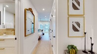 This well-appointed studio is a masterclass in efficient, elegant design. The entry hallway sets a refined tone, featuring light hardwood flooring, recessed lighting, a decorative gold-framed mirror, and curated wall art with matching gold frames — offering an inviting introduction to the space.

The open main area seamlessly blends the sleeping, living, and kitchen zones. The sleeping area is anchored by a generously sized upholstered bed, complemented by a wall-mounted flat-screen TV and a compact wooden desk with a bentwood chair — ideal for work or leisure. A ceiling fan with recessed lighting overhead adds both function and style.

The living area features a neutral loveseat sofa, a round glass coffee table, and a floor lamp, all positioned to take full advantage of the expansive floor-to-ceiling windows that flood the space with natural light and offer views of the surrounding cityscape.

The kitchen is outfitted with white shaker cabinetry, a striking marble-look backsplash, white quartz countertops, brushed gold hardware and faucet, a stainless steel undermount sink, and a full suite of stainless steel appliances.

The bathroom is finished with warm travertine-style tile, a floating white vanity with gold fixtures, and clean, modern lines throughout. A mirrored walk-in closet provides ample storage, completing this compact yet highly functional space.
