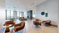 Located at 12 Church Street in New Rochelle, NY, this generously sized amenity floor offers a sophisticated blend of indoor and outdoor spaces designed for comfort, socializing, and relaxation. The interior lounge areas are defined by soaring ceilings, large floor-to-ceiling windows, and polished large-format tile flooring that runs throughout, creating a seamless and airy aesthetic. Curated seating arrangements featuring plush, contemporary chairs and sofas are anchored by circular accent rugs and complemented by striking pendant lighting with gold and matte disc fixtures. A sleek, tile-clad fireplace serves as a dramatic focal point in the main lounge, while floor-to-ceiling windows frame panoramic views of the New Rochelle skyline. A dedicated wet bar and coffee station with warm wood cabinetry provides a functional hub for entertaining. A separate dining and workspace area offers a round table with upholstered seating alongside a built-in counter bar. The space extends outdoors onto multiple terraces, each finished with large-format pavers and outfitted with weather-resistant lounge furniture, offering expansive open-air views of the surrounding urban landscape. Decorative planters and sculptural art elements add character to the outdoor areas, rounding out this polished and well-appointed amenity floor.