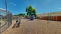 This outdoor playground offers a thoughtfully designed recreational space ideal for children. At the center of the area stands a multi-level play structure complete with a covered canopy top, climbing elements, a colorful slide, and adjacent swing set — providing a variety of engaging activities. The ground is covered in a generous layer of wood chip mulch, offering a cushioned, safety-conscious surface throughout the play area.

The space is fully enclosed by a tall, solid wood privacy fence, creating a secure and defined boundary. Metal gate entries provide controlled access points, connecting the playground to the surrounding property. Additional benches are positioned around the perimeter, offering comfortable seating for supervisors and visitors.

Mature trees within and around the space provide natural shade over portions of the playground, adding a welcoming, green ambiance under open skies. The aerial view of the space reveals a generously sized, triangular footprint that accommodates multiple play zones, including additional smaller play equipment visible toward the far end of the area. The surrounding building and covered parking structure are visible beyond the fencing, situating this playground as an accessible community amenity.