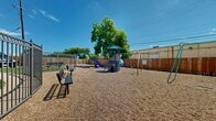 This outdoor playground offers a thoughtfully designed recreational space ideal for children. At the center of the area stands a multi-level play structure complete with a covered canopy top, climbing elements, a colorful slide, and adjacent swing set — providing a variety of engaging activities. The ground is covered in a generous layer of wood chip mulch, offering a cushioned, safety-conscious surface throughout the play area.

The space is fully enclosed by a tall, solid wood privacy fence, creating a secure and defined boundary. Metal gate entries provide controlled access points, connecting the playground to the surrounding property. Additional benches are positioned around the perimeter, offering comfortable seating for supervisors and visitors.

Mature trees within and around the space provide natural shade over portions of the playground, adding a welcoming, green ambiance under open skies. The aerial view of the space reveals a generously sized, triangular footprint that accommodates multiple play zones, including additional smaller play equipment visible toward the far end of the area. The surrounding building and covered parking structure are visible beyond the fencing, situating this playground as an accessible community amenity.
