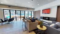 This contemporary single-floor community amenity space is thoughtfully designed to encourage socializing and relaxation. The main lounge area is anchored by a sleek fireplace framed by textured geometric wall panels in warm earth tones, complemented by a wall-mounted flat-screen TV. Plush sectional sofas and coordinated armchairs in neutral and navy tones are arranged to create inviting conversation areas, while accent tables and potted greenery add a fresh, lively touch throughout.

Large floor-to-ceiling sliding glass doors flood the interior with natural light and open directly onto a spacious outdoor terrace, where additional seating is arranged beneath pergola structures — seamlessly blending indoor and outdoor living. A dining area near the terrace access features a solid wood table with modern blue leather chairs, ideal for casual gatherings.

The kitchen is a standout feature, boasting a generous island with a white quartz countertop and bar seating, pendant lighting with amber glass shades, rich wood cabinetry, and a striking shimmering mosaic tile backsplash. A teal accent wall and built-in appliances complete the sophisticated look. Adjacent to the main lounge, a game room with a billiards table provides additional entertainment options, making this space a well-rounded hub for community life.