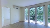 This single-floor space offers a clean, modern aesthetic defined by crisp white walls, smooth ceilings, and light hardwood-style flooring that runs throughout. The main living area is bright and airy, benefiting from an abundance of natural light that pours in through large, floor-to-ceiling glass doors. These doors open directly onto a private balcony enclosed by a sleek metal railing, with mature, leafy trees providing a verdant and tranquil backdrop — a standout feature that brings a sense of nature and privacy to the interior. A wall-mounted split air conditioning unit ensures year-round climate comfort while maintaining a clean, uncluttered look on the walls. The minimalist design of the space allows its architectural elements to take center stage: the generous proportions, the seamless indoor-outdoor connection, and the soft, neutral color palette all work together to create a calm and versatile environment. The interior is well-suited for a variety of uses, offering an open and adaptable layout. Overall, the space strikes a balance between functional modern living and a serene, nature-connected atmosphere.