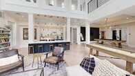 Located at Harpers Crossing in Langhorne, PA, this single-floor communal living space offers a thoughtfully designed, open-concept layout that blends modern farmhouse aesthetics with inviting functionality. The interior is immediately striking, anchored by soaring double-height ceilings that flood the space with natural light through an expansive array of tall, multi-pane windows. White shiplap accent walls add architectural character throughout, complementing the light-toned herringbone hardwood flooring that unifies the entire space.

The main lounge area is generously proportioned, featuring a large sectional sofa, a glass-top coffee table, and a wall-mounted flat-screen TV atop a stylish wood-and-black media console. Lush indoor greenery enhances the fresh, airy atmosphere. Adjacent to the lounge, a bar counter with a dark navy base and pendant globe lighting in brushed gold provides a sophisticated gathering spot, complete with bar stools and open shelving.

The entertainment zone includes a full-size pool table with a contemporary light-wood frame, a shuffleboard table, and a built-in bookcase. A staircase with sleek black metal railings leads to an upper mezzanine level, adding vertical dimension. Curated artwork and decorative wall accents round out the space, creating a polished yet welcoming environment ideal for social gatherings and leisure.