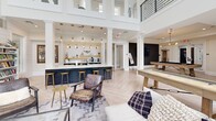 Located at Harpers Crossing in Langhorne, PA, this single-floor communal living space offers a thoughtfully designed, open-concept layout that blends modern farmhouse aesthetics with inviting functionality. The interior is immediately striking, anchored by soaring double-height ceilings that flood the space with natural light through an expansive array of tall, multi-pane windows. White shiplap accent walls add architectural character throughout, complementing the light-toned herringbone hardwood flooring that unifies the entire space.

The main lounge area is generously proportioned, featuring a large sectional sofa, a glass-top coffee table, and a wall-mounted flat-screen TV atop a stylish wood-and-black media console. Lush indoor greenery enhances the fresh, airy atmosphere. Adjacent to the lounge, a bar counter with a dark navy base and pendant globe lighting in brushed gold provides a sophisticated gathering spot, complete with bar stools and open shelving.

The entertainment zone includes a full-size pool table with a contemporary light-wood frame, a shuffleboard table, and a built-in bookcase. A staircase with sleek black metal railings leads to an upper mezzanine level, adding vertical dimension. Curated artwork and decorative wall accents round out the space, creating a polished yet welcoming environment ideal for social gatherings and leisure.