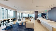 This elegantly designed amenity floor offers a seamless blend of social, recreational, and productive spaces within a modern, high-rise setting. The open-plan layout is anchored by a generously proportioned lounge area furnished with plush, neutral-toned sectional sofas, deep blue area rugs, and distinctive sculptural side tables in bold yellow accents, creating a sophisticated yet inviting atmosphere.

A standout feature is the large communal island bar, finished in crisp white stone and lined with sleek black bar stools, illuminated overhead by striking sputnik-style chandeliers. Floor-to-ceiling glass walls flood this area with abundant natural light and frame panoramic city skyline views, enhancing the sense of openness and elevation.

Adjacent zones include a dedicated co-working nook with built-in cabinetry, a wall-mounted monitor, and a desk workspace, alongside a media lounge featuring a recessed TV wall finished in textured grey paneling with a built-in bench seat. Light herringbone-patterned flooring flows throughout, contrasted by large-format marble-look tile near the pool access corridor.

An expansive wall of windows overlooks the outdoor pool deck, accessible via a raised marble-tiled stairway with black metal railings. Lush potted greenery is thoughtfully placed throughout, adding warmth and vibrancy to the contemporary design. The overall ambiance is polished, airy, and distinctly urban.