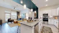 This well-appointed single-floor residence offers a thoughtfully designed open-concept layout that seamlessly connects the kitchen, dining, and living areas. The kitchen is a standout feature, boasting tall white shaker cabinetry, a gray tile backsplash, stainless steel appliances, and a generous island with striking veined stone countertops and pendant lighting overhead — ideal for both cooking and casual dining with barstool seating. Light wood-look flooring runs throughout the main living areas, complementing the bright, airy atmosphere enhanced by elevated ceilings with a bold teal accent. The living room offers large windows with ample natural light and views to the outdoors, creating a warm and inviting atmosphere. Two well-proportioned bedrooms feature plush carpeting, ceiling fans, and large windows. The primary bathroom includes a granite-topped vanity, wood-look tile flooring, and a glass-enclosed shower. A separate powder room with a pedestal sink provides added convenience for guests. Completing the home is a dedicated laundry area with a full-size washer and dryer. The overall design balances modern finishes with comfortable, functional living spaces.