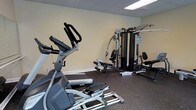 This single-floor fitness room offers a practical and functional space designed to accommodate a variety of workout routines. The room features durable rubber flooring, ideal for supporting heavy equipment and providing a safe surface for exercise. The walls are finished in a neutral, light tone, contributing to a clean and focused environment, while recessed ceiling tiles allow for even overhead lighting throughout the space.

The cardio section is equipped with an elliptical trainer and a treadmill, providing options for low-impact and moderate-intensity cardiovascular workouts. Alongside these machines, a multi-station cable and weight training system anchors the strength training area, offering multiple attachment points and seating configurations to target a wide range of muscle groups. The layout is thoughtfully arranged to allow users to move between stations with ease.

Overall, the space presents a straightforward and well-organized fitness environment. Its compact yet efficient design makes it suitable for individual use, offering both cardio and strength training capabilities within a single, accessible room.