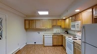 This single-floor space centers around a well-appointed kitchen designed for both practicality and comfort. The kitchen features warm-toned wood cabinetry that lines the upper and lower walls, providing ample storage throughout. Light granite-style countertops complement the cabinetry, offering generous workspace for meal preparation. The layout follows an L-shaped configuration, efficiently organizing the cooking and cleaning zones. A stainless-style sink is positioned at the corner junction, flanked by a white dishwasher and a white electric range with coil burners. A built-in microwave is mounted above the range, maximizing counter space. Overhead, a large flush skylight serves as the kitchen's primary light source, flooding the space with bright, even illumination and lending an open, airy feel to the room. Recessed lighting provides additional ambient light. The flooring consists of light-toned laminate wood planks, which enhance the brightness of the space and tie together the neutral color palette. White baseboards and light-colored walls contribute to the clean, streamlined aesthetic throughout.