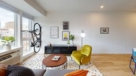 This single-floor space offers a thoughtfully designed open-concept layout that seamlessly connects the living, dining, and kitchen areas into one cohesive environment. Light hardwood flooring runs throughout, contributing to a warm and inviting atmosphere complemented by crisp white walls and recessed lighting.

The kitchen is outfitted with rich dark-wood cabinetry, light quartz countertops, and a geometric tile backsplash. Stainless steel appliances—including a gas range, built-in microwave, and dishwasher—are integrated cleanly into the cabinetry, while an open shelf adds a stylish display element. A large window beside the kitchen allows natural light to pour in, offering views to the outside.

The living area is bright and open, featuring large windows that further enhance the sense of space. The layout comfortably accommodates a seating arrangement alongside a media console, with wall art adding personality to the space.

The sleeping area flows naturally from the main living zone, providing a private yet connected retreat. The bathroom features large-format linear tile in neutral tones lining the tub-shower surround, paired with classic chrome fixtures and hardwood flooring, creating a clean and functional space.

Overall, the unit presents a contemporary, well-appointed interior with cohesive design elements, natural light, and a practical open floor plan ideal for modern living.