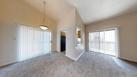 Located at Sun Creek on Spain Road NE in Albuquerque, New Mexico, this single-floor residence showcases an open and airy layout designed to maximize comfort and functionality. The living and dining area immediately impresses with its dramatic vaulted ceilings, which lend the space a sense of generous volume and openness. Plush wall-to-wall carpeting in a neutral tone runs throughout, providing a warm and cohesive feel underfoot. Large sliding glass doors fitted with vertical blinds flood the interior with natural light while offering direct access to the outdoors and views of the surrounding exterior. A stylish pendant light fixture anchors the dining area, adding a subtle decorative touch to the open-plan space. A half-wall partition elegantly separates the living area from the kitchen pass-through, maintaining visual connectivity while defining each zone. The kitchen, visible through the pass-through opening, features cabinetry and standard appliances, including a full-size refrigerator. The interior's neutral color palette of soft whites and warm beige tones creates a clean, versatile backdrop well-suited to a variety of furnishing styles. Overall, the space presents a bright, well-proportioned single-level layout that balances architectural character with everyday practicality.