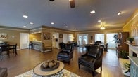 This single-floor space is designed with an open-concept layout that seamlessly connects multiple functional areas, creating a welcoming and versatile environment. The main living area is anchored by rich hardwood flooring and features a seating arrangement with dark leather armchairs accented by nailhead trim, centered around a round coffee table with decorative accents. A patterned area rug adds visual warmth beneath the furniture grouping.

Recessed lighting is installed throughout the ceiling, providing consistent illumination across the space, while ceiling fans contribute to both comfort and airflow. The walls are finished in warm earth tones, complemented by white crown molding that defines the transitions between areas.

Toward the rear, French doors flood the space with natural light and offer a view to the exterior. A dedicated work or meeting area with a desk and seating is visible nearby. The kitchen or kitchenette area, partially separated by a half-wall partition, includes bar-height seating along a counter, cabinetry, and appliances. Decorative wall art and subtle fixtures complete the polished, cohesive aesthetic of this well-appointed space.