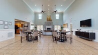 This single-story community clubhouse is thoughtfully designed to serve as a welcoming hub for residents and guests alike. The centerpiece is a grand great room boasting soaring double-height ceilings, soft sage-blue walls accented by crisp white wainscoting, and large tile flooring throughout. Dual ceiling fans maintain comfort, while an array of tall windows and French doors flood the space with natural light and provide views of the surrounding greenery. A central fireplace anchors the room, which is furnished with multiple high-top dining tables and chairs, making it ideal for casual gatherings. Adjacent to the great room, a comfortable lounge area features leather seating, wall-mounted artwork, and a flat-screen television, offering a relaxed retreat. A dedicated media room provides an intimate home-theater setup with plush seating, wall-mounted speakers, and a large-screen TV. The fully equipped kitchen includes warm wood cabinetry, a mosaic tile backsplash, a center island, and full-size appliances. A professional leasing office with a reception desk, display shelving, and natural light completes the space. The welcoming entry foyer, finished with tile flooring and bright white French doors, sets an inviting tone throughout.