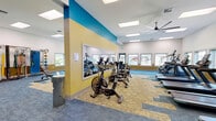 Located at 3280 SW 170th Ave in Beaverton, Oregon, this single-floor fitness center offers a spacious and thoughtfully designed exercise environment. The room is characterized by soaring ceilings, large skylights, and rows of windows along the walls that flood the space with natural light and provide views of the surrounding outdoors. A large ceiling fan contributes to comfortable airflow throughout.

The interior design features a bold, energizing color palette with teal and golden-yellow accent walls accented by full-length mirrors, all set against a patterned carpet tile flooring in shades of blue and tan. The cardio zone is well-appointed with multiple treadmills, stationary bikes, and elliptical machines arranged in rows for easy access. A dedicated strength training area includes adjustable weight benches, kettlebell racks, and cable machines, while a functional training zone along one wall is equipped with Smart Trainer racks, TRX suspension straps, resistance bands, battle ropes, and a variety of stability and medicine balls. The layout provides generous open floor space for stretching and movement, making it suitable for a wide range of workout styles and fitness levels.