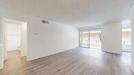 Located on Sherman Way in the Reseda neighborhood of Los Angeles, this one-bedroom, one-bathroom unit offers a well-organized layout across a single floor. Light wood-style flooring flows consistently throughout the space, lending a clean, cohesive aesthetic to every room.

The living and dining areas are open and connected, bathed in natural light from a full-width sliding glass door that leads to a private balcony. The vaulted ceiling detail in the living area adds a sense of vertical space, while a ceiling fan provides year-round comfort. The balcony offers outdoor access with metal railing, reflective of the building's multi-story stucco exterior.

The galley-style kitchen is well-appointed with white cabinetry featuring matte black hardware, light countertops, a stainless steel refrigerator, and a white range. Overhead lighting illuminates the full length of the kitchen, which flows toward the dining area.

The bedroom is generously sized with a large window admitting natural light, and benefits from ample closet storage — the unit includes four closets in total. A hallway connects the main rooms efficiently.

The bathroom is clean and modern, featuring a white vanity with black hardware, a mirror, and wood-style flooring that matches the rest of the unit. The textured ceilings throughout give the space a consistent architectural character.