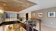 This two-floor space offers a generously sized lower level and a more intimate upper mezzanine, connected by a striking open staircase with sleek dark metal cable railings. The lower level serves multiple functions, beginning with a dedicated bike storage and maintenance room featuring polished concrete floors, wall-mounted bike racks, vibrant cycling-themed murals, a repair station, and cushioned benches. Adjacent areas include a relaxed lounge nook with suspended rattan hanging chairs, live-edge wood stump stools, white brick accent walls, and warm pendant lighting beneath a reclaimed wood ceiling detail.

The main lobby area is anchored by the open staircase and features an elevator with white brick surround, framed artwork, and polished concrete flooring throughout. A large-scale nature mural and playful illustrated wall art add personality and visual interest.

The upper mezzanine level overlooks the main floor through the open railing system and is characterized by reclaimed wood plank ceiling panels, recessed lighting, neutral toned walls, potted greenery, and framed art displays. The overall design aesthetic combines industrial elements—dark metal, polished concrete—with organic warmth through wood, plants, and artful, community-oriented décor.