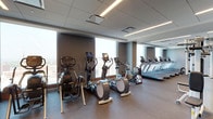 The Omni fitness center is a spacious, contemporary facility designed to accommodate a comprehensive workout experience. The space features a clean, modern aesthetic defined by a neutral gray color palette, drop-panel ceilings with integrated linear lighting, and durable rubberized flooring throughout.

The main cardio area is anchored by a row of commercial-grade treadmills equipped with individual screens, complemented by an array of elliptical trainers and stair climbers. Floor-to-ceiling windows line the exterior wall, flooding the space with natural light and offering sweeping elevated views of the surrounding cityscape, providing an energizing backdrop during workouts.

A dedicated strength training zone features cable machines, weight benches, and a full dumbbell rack, all reflected by mirrored walls that visually expand the room. A separate functional training area includes Concept2 rowing machines and stability balls stored on built-in wooden shelving units, offering a more intimate setting for cross-training.

A wall-mounted flat-screen television is positioned in the cardio section for entertainment. The overall ambiance is polished and professional, combining high-quality equipment with thoughtful design to deliver a premium fitness environment.