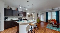 This single-floor residence offers a thoughtfully designed open-concept layout that seamlessly connects the kitchen, dining, and living areas. Rich hardwood-style flooring runs throughout the main living spaces, complemented by crown molding and ceiling fans that add a polished finish.

The kitchen is a standout feature, equipped with dark espresso cabinetry, stainless steel appliances including a refrigerator, range, and microwave, and granite countertops. A curved breakfast bar with pendant lighting above provides both additional prep space and casual seating. Track lighting overhead ensures the kitchen is well-lit for cooking and entertaining.

The open living area is bright and inviting, with a ceiling fan and access to a glazed entry door framed by teal curtains that allow natural light to filter in. The space flows naturally into a defined dining area, making the layout highly functional.

The bedroom is a calm retreat, featuring a ceiling fan, carpeted flooring, and a window with ample natural light. It connects directly to a generous walk-in closet outfitted with multi-level shelving and hanging rods, offering excellent storage capacity.

The bathroom is finished with tile flooring, a granite-topped vanity with dark cabinetry, and a full tub-shower combination with tile surround. Clean, neutral finishes throughout create a cohesive and comfortable living environment.
