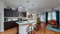 This single-floor residence offers a thoughtfully designed open-concept layout that seamlessly connects the kitchen, dining, and living areas. Rich hardwood-style flooring runs throughout the main living spaces, complemented by crown molding and ceiling fans that add a polished finish.

The kitchen is a standout feature, equipped with dark espresso cabinetry, stainless steel appliances including a refrigerator, range, and microwave, and granite countertops. A curved breakfast bar with pendant lighting above provides both additional prep space and casual seating. Track lighting overhead ensures the kitchen is well-lit for cooking and entertaining.

The open living area is bright and inviting, with a ceiling fan and access to a glazed entry door framed by teal curtains that allow natural light to filter in. The space flows naturally into a defined dining area, making the layout highly functional.

The bedroom is a calm retreat, featuring a ceiling fan, carpeted flooring, and a window with ample natural light. It connects directly to a generous walk-in closet outfitted with multi-level shelving and hanging rods, offering excellent storage capacity.

The bathroom is finished with tile flooring, a granite-topped vanity with dark cabinetry, and a full tub-shower combination with tile surround. Clean, neutral finishes throughout create a cohesive and comfortable living environment.
