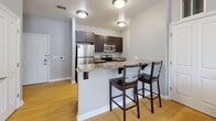 Located at 18490 Dunn Rd in South Bend, Indiana, this single-floor, 2-bedroom, 2-bathroom residence offers a thoughtfully designed layout that makes efficient use of its space. The kitchen is a standout feature, showcasing rich dark cabinetry paired with granite-style countertops, stainless steel appliances including a refrigerator, microwave, and range, and warm hardwood-style flooring. A peninsula with an overhanging counter and bar seating for two creates a casual dining area that connects seamlessly to the living space. Both bedrooms are carpeted with patterned flooring and feature neutral gray walls that lend a calm, modern ambiance. Each room benefits from natural light provided by windows fitted with horizontal blinds. The primary bedroom offers a cozy, functional layout with room for standard furnishings. The two bathrooms are finished with large-format tile flooring, dark vanity cabinetry with white countertops, and tub/shower combinations, providing clean and practical design. The overall aesthetic throughout the residence is contemporary and neutral, with consistent use of gray tones, white trim, and dark wood accents creating a cohesive and inviting living environment.