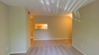This single-story space presents a straightforward and functional layout centered around a spacious living area finished with wall-to-wall carpeting in a neutral beige tone. The room features clean white baseboards and smooth, light-colored walls that contribute to a warm and understated ambiance. A distinctive architectural detail is the pass-through window cut into the wall, which visually connects the living area to the kitchen, promoting an open feel while maintaining a defined separation between spaces. Through the pass-through, the kitchen is visible and features wood-toned cabinetry, a countertop with what appears to be a stone or laminate surface, and under-cabinet lighting that casts a warm glow. A hallway is visible to the left, suggesting additional rooms beyond the main living space. The ceiling includes a textured or paneled section near the front of the room, adding a subtle design element overhead. Overhead lighting provides consistent illumination throughout. The overall atmosphere is warm and neutral, offering a blank canvas that is well-suited for a variety of furnishing styles.