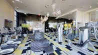 This fitness center makes an immediate impression with its fearlessly creative interior design. The space is anchored by a bold, graphic-patterned floor in black, white, and yellow that sets an energetic tone throughout. Overhead, a dramatic cluster of teardrop-shaped pendant lights in black-and-white zebra stripes draws the eye upward and serves as a stunning focal point. The dark, high-gloss accent walls contrast sharply with the lighter ceiling, lending the space a sleek, modern edge. A large-scale graphic mural adds an artistic, editorial quality to the room. Floor-to-ceiling mirrors expand the sense of space and allow for form-checking during workouts. The fitness center is well-equipped with a range of strength and cardio machines, along with an assortment of colorful medicine balls and free weights neatly arranged for easy access. A lounge chair with a modern, tufted design offers a place to rest between sets, blurring the line between gym and lifestyle space. Mounted flat-screen televisions provide entertainment. The overall ambiance is that of a high-end, design-forward fitness facility that is as visually stimulating as it is functional.