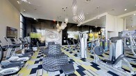 This fitness center makes an immediate impression with its fearlessly creative interior design. The space is anchored by a bold, graphic-patterned floor in black, white, and yellow that sets an energetic tone throughout. Overhead, a dramatic cluster of teardrop-shaped pendant lights in black-and-white zebra stripes draws the eye upward and serves as a stunning focal point. The dark, high-gloss accent walls contrast sharply with the lighter ceiling, lending the space a sleek, modern edge. A large-scale graphic mural adds an artistic, editorial quality to the room. Floor-to-ceiling mirrors expand the sense of space and allow for form-checking during workouts. The fitness center is well-equipped with a range of strength and cardio machines, along with an assortment of colorful medicine balls and free weights neatly arranged for easy access. A lounge chair with a modern, tufted design offers a place to rest between sets, blurring the line between gym and lifestyle space. Mounted flat-screen televisions provide entertainment. The overall ambiance is that of a high-end, design-forward fitness facility that is as visually stimulating as it is functional.