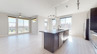 This single-floor residence showcases a thoughtfully designed open-concept layout bathed in natural light. The living and dining area flows seamlessly from the kitchen, featuring light hardwood-style flooring, track lighting, and large floor-to-ceiling windows that frame sweeping urban skyline views. A balcony access door extends the living space outdoors.

The kitchen is a standout, centered around a substantial island with a dark wood finish, integrated sink, and a sleek pendant light fixture overhead. Cabinetry along the perimeter features a gray wood-grain finish, complemented by a decorative subway tile backsplash with subtle color accents. Stainless steel appliances — including a refrigerator, electric range, microwave, and dishwasher — complete the functional space.

The bedroom is generously sized with plush carpeting, a ceiling fan, and dual large windows offering bright city views. Two bathrooms serve the residence: one features a soaking tub with full floor-to-ceiling tile surround, while the other offers a long vanity with dark cabinetry and stone-look countertops, with light tile flooring throughout both. The overall aesthetic is clean, contemporary, and urban.