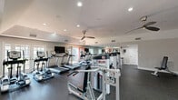 Located in Palm Beach Gardens, FL, this fitness center offers a spacious and thoughtfully organized workout facility spread across a single floor. The space is divided into two dedicated exercise rooms, each designed to accommodate a variety of fitness routines and training styles.

The main cardio area features a row of TRUE treadmills and elliptical machines aligned along the room, complemented by large wall mirrors that visually expand the space and allow users to monitor their form. Ceiling fans provide comfortable airflow throughout, while recessed lighting ensures a bright, energizing atmosphere. French-style doors with glass panels invite natural light and offer views to the outdoors.

The strength training area is equipped with a Smith machine, free weights, adjustable benches, and a cable resistance system, providing ample options for full-body workouts. Rubber flooring throughout both rooms ensures durability and safety. A water bottle refill station and waste receptacle are conveniently positioned near the entrance, adding a practical amenity for users.

The clean, neutral-toned walls and efficient layout create an inviting and functional environment suitable for a wide range of fitness levels. The use of mirrors, ceiling fans, and natural light through multiple windows contributes to an open, well-ventilated space that supports an effective and comfortable workout experience.