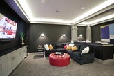 This stylish single-floor entertainment lounge is designed with a refined, contemporary aesthetic that balances bold design choices with comfortable functionality. The space is defined by dramatic dark houndstooth-patterned wall treatments that create a rich, moody atmosphere, complemented by crisp white architectural columns and detailed millwork that add a classic contrast.

Cove lighting runs along the perimeter of a coffered ceiling, casting a warm, ambient glow throughout the room. Recessed and wall-mounted sconces provide additional layered lighting, enhancing the sophisticated ambiance. The flooring features a plaid-patterned carpet in muted tones, adding texture while grounding the space.

A central highlight is the media area, where a large wall-mounted flat-screen television is anchored above a row of white built-in cabinetry with gold hardware and a dark countertop, ideal for housing media equipment and display pieces. A sleek bar counter with white bar stools serves as both a functional and social focal point, separating zones within the space.

The lounge seating area includes a deep navy sectional sofa accented with vibrant throw pillows and a tufted red ottoman, creating a lively and inviting gathering spot. A pair of cognac leather armchairs offers additional seating in a quieter corner. Bold geometric artwork adorns the walls, adding a curated, gallery-like quality to the overall design.