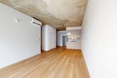 Located at 2600 Cavendish Boulevard in Montréal, this two-bedroom, one-bathroom unit offers a clean, contemporary aesthetic defined by a striking combination of exposed concrete ceilings and warm light hardwood flooring throughout. The open-plan living and kitchen area forms the heart of the unit, creating a spacious, airy atmosphere enhanced by crisp white walls and a wall-mounted air conditioning unit. The kitchen is well-appointed with a central island featuring a white quartz countertop and an undermount sink, complemented by light wood cabinetry, white upper cabinets, stainless steel appliances including a refrigerator and dishwasher, and a freestanding range. A pendant light fixture adds a refined finishing touch. The living area benefits from a large window overlooking the surrounding urban streetscape, filling the space with natural light. Both bedrooms continue the hardwood flooring and exposed concrete ceiling motif, maintaining the unit's cohesive industrial-modern character. The primary bedroom includes a built-in closet, while the second bedroom also features a window with city views. The bathroom is finished in a minimalist style with large-format light tile flooring, a floating vanity, a bathtub with tiled surround, and recessed lighting.