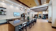 This single-floor community amenity space is thoughtfully designed to offer both function and comfort through a series of well-appointed areas. The entrance corridor features light wood-toned flooring and clean white walls, leading into a grand reception area with soaring ceilings, polished tile floors, and large windows that invite natural light. The central gathering area is anchored by a substantial dark granite island bar surrounded by upholstered counter-height chairs, illuminated by striking sculptural pendant chandeliers with an intertwining metallic design. Exposed dark wood ceiling beams add warmth and architectural character overhead. The kitchenette runs along one wall and features sleek dark wood cabinetry, a stainless steel oven, white countertops, an undermount sink, wall-mounted television, and a Starbucks coffee station — making it a fully equipped refreshment hub. A separate lounge area offers an intimate retreat with rich emerald green velvet sofas, a tufted upholstered accent wall, textured wall paneling, a fireplace, and large windows overlooking an outdoor terrace. Abstract wall art and carefully curated décor add a residential sophistication throughout. The light hardwood flooring and neutral palette create a cohesive, welcoming ambiance across the entire space.