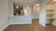 This single-floor space offers a clean, contemporary design centered around an open-concept living and kitchen area. The kitchen is a standout feature, boasting a large white waterfall-edge island with an undermount sink, dark cabinetry, stainless steel appliances including a French door refrigerator and built-in microwave, and a sleek tile backsplash. Pendant lighting with glass dome shades hangs above the island, complemented by track lighting overhead, ensuring the space is well-lit and visually inviting. The flooring throughout is a warm hardwood-style material that adds texture and cohesion to the open layout. Neutral gray walls provide a versatile backdrop that suits a variety of interior styles. The living area flows naturally from the kitchen, creating an expansive, connected feel. A hallway leads to additional rooms, with clean white doors and crisp white baseboards maintaining the modern aesthetic. The overall ambiance is bright, airy, and move-in ready, combining functional design with contemporary finishes that appeal to those seeking a polished, low-maintenance living environment.