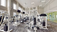 This single-floor fitness facility is designed with both form and function in mind, combining classic architectural elegance with a comprehensive array of workout amenities. The main gym area commands attention with soaring double-height ceilings, large arched windows that flood the space with natural light, and ceiling fans that maintain a comfortable atmosphere. Wainscoting and paneled walls in a neutral palette give the space a refined, upscale character. The gym floor is finished with a durable multi-tone carpet tile and is well-stocked with strength training machines, cable systems, a full dumbbell rack, treadmills, ellipticals, and stationary bikes, all complemented by wall-mounted televisions. Full-length mirrors along the walls enhance the sense of openness and allow users to monitor their form. Adjacent to the main workout area, a dedicated racquetball court features a polished hardwood floor with regulation court markings, crisp white walls, and recessed overhead lighting. A connecting corridor provides access to water fountains, vending machines, and additional fitness amenities, ensuring convenience throughout. Direct exterior access connects the facility to outdoor areas, making this a fully self-contained and thoughtfully appointed athletic destination.