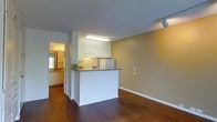 This single-floor unit features an open-concept layout that connects the kitchen and living area, creating a functional and cohesive living space. The kitchen is equipped with white shaker-style cabinetry, granite countertops, and a peninsula that serves as both a workspace and a divider between the kitchen and the adjoining living area. A fluorescent light fixture provides direct illumination over the kitchen workspace, while a track light in the living area offers additional lighting flexibility. The flooring throughout is a warm-toned hardwood, adding character and continuity across the space. Walls are painted in a neutral tan hue, complementing the white trim and cabinetry. A textured popcorn ceiling adds a classic touch overhead. Visible through a doorway is an additional room — likely a bathroom or vanity area — featuring wood-toned cabinetry, a lighted mirror, and a window that allows natural light to filter in. The overall design is straightforward and practical, with clean finishes and a neutral palette that offers a versatile backdrop for various interior styles.