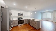 Located at 3857 Pechin Street in Philadelphia's Roxborough neighborhood, this well-appointed one-bedroom, one-bathroom residence offers a thoughtfully designed single-floor layout. The open-concept kitchen is a standout feature, showcasing a two-tone cabinet design with crisp white upper cabinets and textured gray lower cabinets, complemented by white quartz countertops and a peninsula with pendant lighting. Stainless steel appliances, including a range, microwave, dishwasher, and refrigerator, complete this modern kitchen, which flows seamlessly into the living area with rich hardwood flooring throughout. The bedroom is a comfortable, carpeted retreat featuring a wide window with views of greenery, filling the room with natural light. A dedicated closet provides practical storage. The bathroom is finished with large-format marble-look floor tiles, a sleek glass-enclosed walk-in shower with a rainfall showerhead, and clean white walls that create a bright, spa-like atmosphere. A dedicated laundry area houses a stacked full-size washer and dryer, adding everyday convenience. The overall design reflects a clean, modern aesthetic with neutral tones and quality finishes throughout.