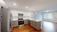 Located at 3857 Pechin Street in Philadelphia's Roxborough neighborhood, this well-appointed one-bedroom, one-bathroom residence offers a thoughtfully designed single-floor layout. The open-concept kitchen is a standout feature, showcasing a two-tone cabinet design with crisp white upper cabinets and textured gray lower cabinets, complemented by white quartz countertops and a peninsula with pendant lighting. Stainless steel appliances, including a range, microwave, dishwasher, and refrigerator, complete this modern kitchen, which flows seamlessly into the living area with rich hardwood flooring throughout. The bedroom is a comfortable, carpeted retreat featuring a wide window with views of greenery, filling the room with natural light. A dedicated closet provides practical storage. The bathroom is finished with large-format marble-look floor tiles, a sleek glass-enclosed walk-in shower with a rainfall showerhead, and clean white walls that create a bright, spa-like atmosphere. A dedicated laundry area houses a stacked full-size washer and dryer, adding everyday convenience. The overall design reflects a clean, modern aesthetic with neutral tones and quality finishes throughout.