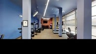 This fitness center offers a comprehensive workout environment designed to accommodate a wide range of exercise needs. The space is characterized by bold blue and terracotta accent walls paired with neutral-toned carpeting, creating an energetic yet focused atmosphere. Sleek linear pendant lighting runs across the ceiling alongside recessed fixtures, ensuring the entire floor is well-illuminated throughout.

The cardio section lines the perimeter near a expansive floor-to-ceiling window wall, where elliptical trainers and treadmills are positioned to take advantage of natural light and outdoor views. The strength training area features multiple cable and resistance machines, a leg extension and curl station, and a dedicated free weight zone stocked with a full rack of dumbbells. Exercise balls in various sizes round out the functional fitness options.

Structural columns divide the space into distinct zones without disrupting the open, airy feel of the floor plan. A wall-mounted flat-screen television is visible from the strength training area, adding an entertainment element to workouts. Double glass doors at the far end provide convenient access and contribute additional natural light to this thoughtfully arranged fitness facility.