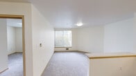 Located at 6940 37th Ave S in Seattle's Brighton neighborhood, this one-bedroom, one-bathroom unit offers a practical and efficiently designed single-floor layout. The open-concept living area features neutral-toned carpet and a large window that invites natural light while providing views of surrounding greenery, creating a calm and airy atmosphere. A half-wall peninsula separates the living space from the kitchen, which is equipped with light maple cabinetry, a white electric range, refrigerator, microwave, and a single-basin sink. The kitchen floor is finished with wood-look vinyl planking, providing a clean contrast to the carpeted areas. The bedroom is a comfortable, well-proportioned room with plush carpet, a built-in closet with a light wood bi-fold door, and floor-level heating ventilation. Its neutral walls and simple layout make it an easily adaptable space. The bathroom is finished with wood-look flooring and features a shower-tub combination, a vanity with countertop sink, and thoughtfully installed grab bars, reflecting an accessibility-conscious design. Throughout the unit, light wood trim accents, flush-mount ceiling lighting, and a clean, neutral color palette contribute to a cohesive and functional living environment.
