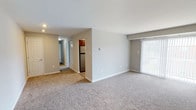 This single-floor unit is designed with a practical, straightforward layout that maximizes usable living space. The open living area is finished with neutral-toned wall-to-wall carpeting and light gray walls, creating a clean and versatile backdrop. A large sliding glass door fitted with vertical blinds allows natural light to fill the space while providing access to the outdoors. The kitchen is efficiently arranged and features rich, warm-toned cabinetry, granite-style countertops, stainless steel appliances including a gas range, over-the-range microwave, and refrigerator, along with a stainless steel sink positioned beneath a window. The bedrooms are carpeted and finished in the same neutral gray palette, each featuring windows with blinds that allow natural light in. One bedroom includes an adjoining closet with warm interior lighting. A hallway connects the living areas to the private spaces, including a bathroom equipped with a tub and shower combination. Throughout the unit, clean white baseboards, recessed lighting, and consistent neutral finishes contribute to a cohesive and well-maintained living environment.