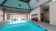 Located at 3751 Appian Way in Lexington, Kentucky, this single-story indoor pool facility offers a well-appointed aquatic space designed for year-round use. The centerpiece is a generously sized indoor swimming pool with clear, blue-tinted water and a tiled surround, providing ample space for lap swimming and leisure alike. The pool area is crowned by striking vaulted ceilings with exposed beams and a teal-painted paneled ceiling, lending the space an open, airy atmosphere. Large glass doors and windows along one wall allow abundant natural light to pour in, offering views of the outdoor surroundings and creating a bright, welcoming environment. The pool deck features durable concrete flooring and is furnished with a mix of seating options, including wicker lounge chairs with bold red cushions and a casual dining table with chairs, ideal for relaxing poolside. A dedicated lounge area with red upholstered seating and a woven coffee table provides a comfortable retreat for those not in the water. Signage throughout ensures safety compliance, including clearly posted pool rules and no-diving notices. Additional utility areas and access doors are conveniently situated around the perimeter, supporting the facility's functionality. The overall ambiance is clean, practical, and inviting, making this a standout community amenity.