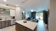 This two-bedroom residence at Tamarac Village showcases a contemporary open-concept design on a single floor. The kitchen is a standout feature, equipped with stainless steel appliances including a refrigerator, range, microwave, and dishwasher. Two-tone cabinetry pairs white upper cabinets with wood-finish lower cabinets, complemented by subway tile backsplash and light quartz countertops. A central island provides additional prep space and casual seating. The kitchen flows seamlessly into the dining and living areas, where light wood-style flooring, a ceiling fan, and sliding glass doors opening to an outdoor patio create a bright, airy atmosphere with natural light.

Both bedrooms feature plush carpeting, warm neutral tones, and generous windows dressed with blinds and floor-length curtains. The primary bedroom benefits from dual windows and a ceiling fan. Two bathrooms serve the unit: one features a glass-enclosed walk-in shower with subway tile surround and a wide vanity, while the second includes a soaking tub/shower combination with matching tile work and a wood-finish vanity. Track and pendant lighting throughout adds a polished, modern touch to the overall design.