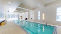 This sophisticated indoor pool facility offers a refined aquatic environment defined by clean lines and a calm, minimalist aesthetic. The centerpiece is a generously sized lap pool with crystal-clear turquoise water, framed by smooth, light-toned tile surrounds that extend across the floor and pool deck. Crisp white walls and a structured ceiling with prominent architectural beams lend the space a sense of height and openness. Tall, narrow windows are evenly spaced along the walls, allowing natural light to filter in and complement the sleek vertical lighting fixtures mounted between them, casting a warm, even glow throughout. At one end, expansive glazed doors and windows open to views of the surrounding urban skyline, further enhancing the bright, airy atmosphere. Stainless steel pool entry rails provide safe and easy access to the water. Thoughtful touches such as potted tropical plants near the entrance add a subtle organic warmth to the otherwise contemporary setting. A row of understated seating is available poolside, providing a place to rest and take in the tranquil surroundings. The overall ambiance is one of quiet luxury—clean, well-appointed, and purpose-built for both fitness and relaxation.