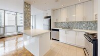 This thoughtfully designed 2-bedroom, 1-bathroom residence offers a functional and stylish layout across a single floor. The open-concept living and dining area is a standout feature, showcasing gleaming parquet hardwood floors throughout and expansive floor-to-ceiling windows that flood the space with natural light while framing impressive urban skyline views. A private balcony extends from the living area, offering an outdoor retreat with sweeping city vistas.

The kitchen is modern and well-equipped, featuring white gloss cabinetry, a decorative mosaic tile backsplash, a breakfast bar island, light tile flooring, stainless steel appliances including a refrigerator and dishwasher, and recessed lighting overhead. A distinctive mosaic tile accent column adds a unique architectural touch at the kitchen's entrance.

Both bedrooms continue the parquet hardwood flooring and are bright, featuring windows with city views, baseboard heating, and double-door closets providing ample storage. The primary bedroom is generously sized with comfortable proportions.

The bathroom is elegantly finished with floor-to-ceiling neutral stone-look tile, a soaking tub with an overhead rain showerhead, a white vanity with a stone countertop, and a large mirror. Clean lines and a contemporary palette define the overall ambiance of this well-appointed residence.