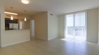 This single-floor space offers an open-concept layout that connects the living area and kitchen in a clean, functional design. The living area is bathed in natural light, courtesy of expansive floor-to-ceiling sliding glass doors fitted with vertical blinds, which open onto a private balcony with views of the surrounding area. Light streams across the neutral-toned ceramic tile flooring, enhancing the airy, spacious feel of the room.

The kitchen is partially visible from the main living space, separated by a partial wall with a pass-through counter, creating a semi-open connection between the two areas. A pendant light hangs above the kitchen counter, adding a subtle decorative touch. The kitchen itself appears compact yet practical.

Throughout the space, smooth white ceilings with recessed ventilation and flush lighting fixtures maintain a clean, modern aesthetic. Neutral beige walls complement the light tile floors, contributing to a cohesive, understated palette. White baseboards frame each room neatly. Overall, the space presents a bright, well-lit environment with an emphasis on openness, natural light, and simple, contemporary finishes.