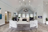 This elegantly designed single-story office space makes an immediate impression with its soaring vaulted ceilings and refined contemporary aesthetic. The centerpiece of the main reception area is a striking curved desk clad in white and grey veined marble, positioned beneath a sculptural sputnik-style chandelier that adds a modern focal point to the vaulted ceiling above. Adjacent to the reception desk, a floor-to-ceiling marble accent wall frames a sleek linear fireplace, lending warmth and a luxurious touch to the space.

The open layout transitions seamlessly into a welcoming lounge and meeting area, furnished with round marble-top tables and tufted grey velvet chairs with chrome frames — a cohesive palette of soft greys, whites, and muted blues throughout. Bold abstract artwork adorns the walls, adding visual interest and a contemporary character.

Expansive floor-to-ceiling windows flood the interior with natural light and offer direct views of an outdoor pool area, blurring the line between inside and out. Rich hardwood-style flooring anchors the space, complemented by layered area rugs with an abstract pattern. A private office is accessible from the main floor, while frosted glass interior doors provide privacy without sacrificing light. The overall ambiance is polished, professional, and inviting.