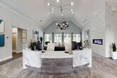 This elegantly designed single-story office space makes an immediate impression with its soaring vaulted ceilings and refined contemporary aesthetic. The centerpiece of the main reception area is a striking curved desk clad in white and grey veined marble, positioned beneath a sculptural sputnik-style chandelier that adds a modern focal point to the vaulted ceiling above. Adjacent to the reception desk, a floor-to-ceiling marble accent wall frames a sleek linear fireplace, lending warmth and a luxurious touch to the space.

The open layout transitions seamlessly into a welcoming lounge and meeting area, furnished with round marble-top tables and tufted grey velvet chairs with chrome frames — a cohesive palette of soft greys, whites, and muted blues throughout. Bold abstract artwork adorns the walls, adding visual interest and a contemporary character.

Expansive floor-to-ceiling windows flood the interior with natural light and offer direct views of an outdoor pool area, blurring the line between inside and out. Rich hardwood-style flooring anchors the space, complemented by layered area rugs with an abstract pattern. A private office is accessible from the main floor, while frosted glass interior doors provide privacy without sacrificing light. The overall ambiance is polished, professional, and inviting.