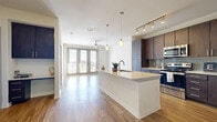 This thoughtfully designed one-bedroom, one-bathroom residence offers a functional and stylish single-floor layout. The open-concept kitchen and living area serves as the heart of the space, where a large center island with granite countertops and an undermount sink takes center stage. The kitchen is equipped with stainless steel appliances, rich dark-toned cabinetry reaching to the ceiling, a herringbone-patterned tile backsplash, and track lighting overhead. The adjoining living room features light hardwood flooring, a ceiling fan, and a set of French doors that invite ample natural light and provide views of the surrounding area. The bedroom is a comfortable retreat with plush carpeting, a window with exterior views, and direct access to the full bathroom. The bathroom is well-appointed with a dual-sink vanity featuring bold blue cabinetry, granite countertops, and large-format tile flooring. A dedicated laundry room with an in-unit washer and additional storage shelving adds everyday practicality. Throughout, the space maintains a clean, contemporary aesthetic with neutral white walls and consistent modern finishes.